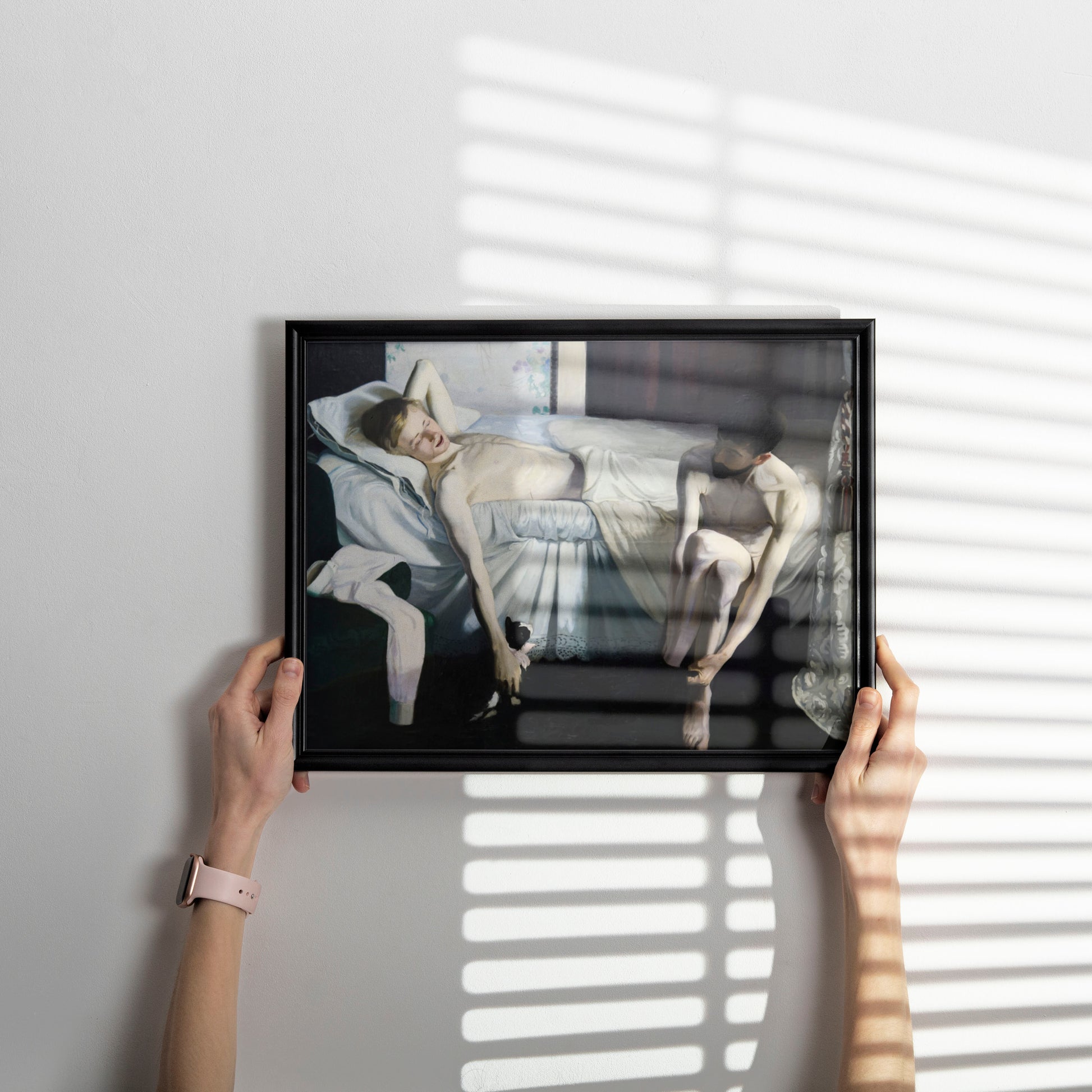 A person holds a black-framed art print against a light gray wall, with soft window-blind shadows cast across the scene. The artwork depicts a reclining nude figure in pale skin tones and white bedding.