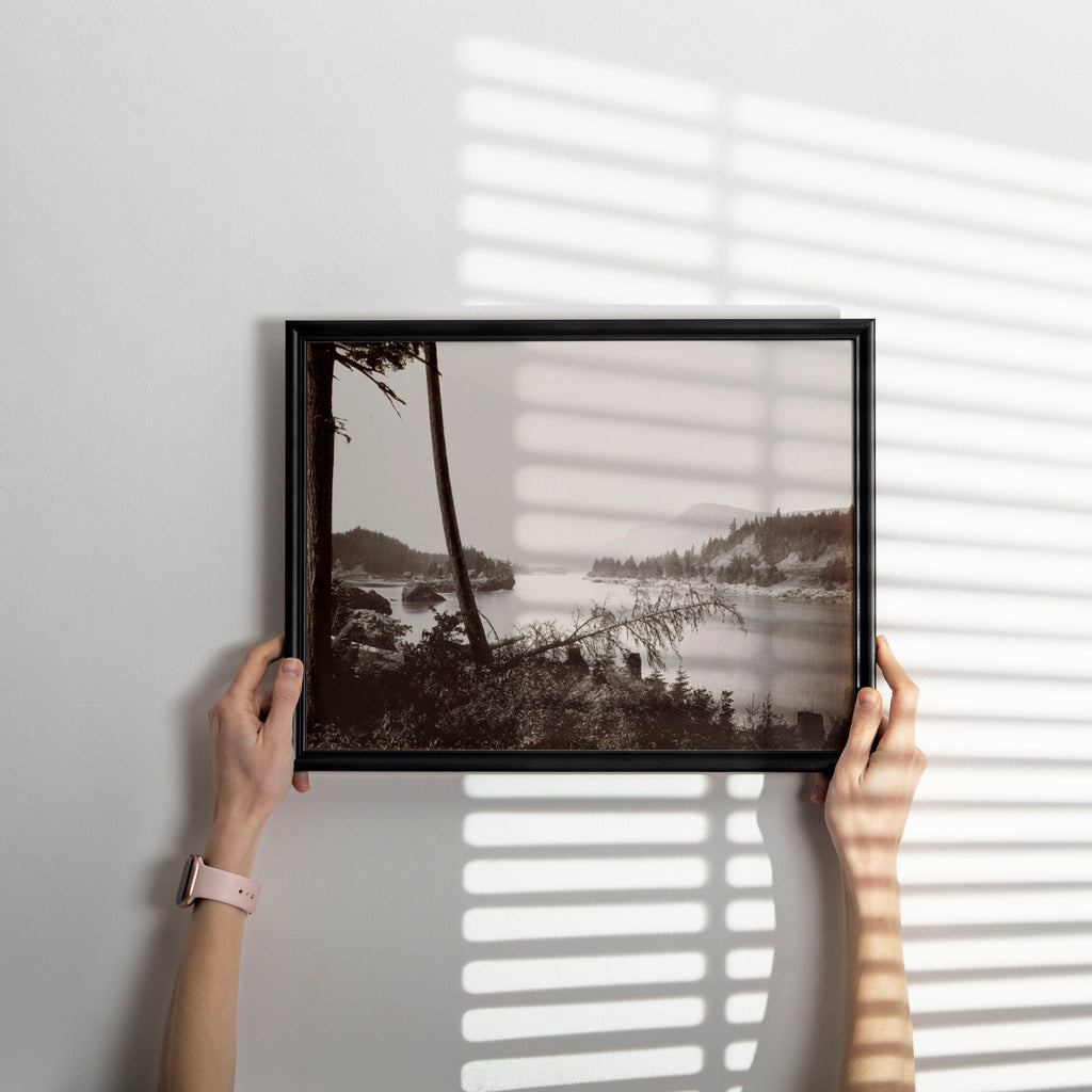 A person holds a black-framed landscape photograph print against a white wall, with soft window-blind shadows cast across the scene.