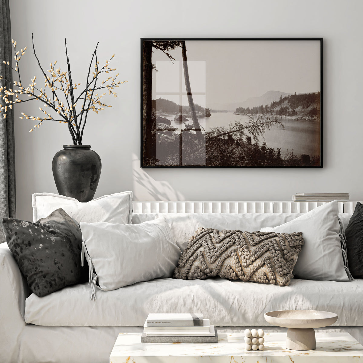A large framed black and white landscape art print hangs on a light gray wall above a white sofa styled with neutral-toned pillows and a dark vase with branches.