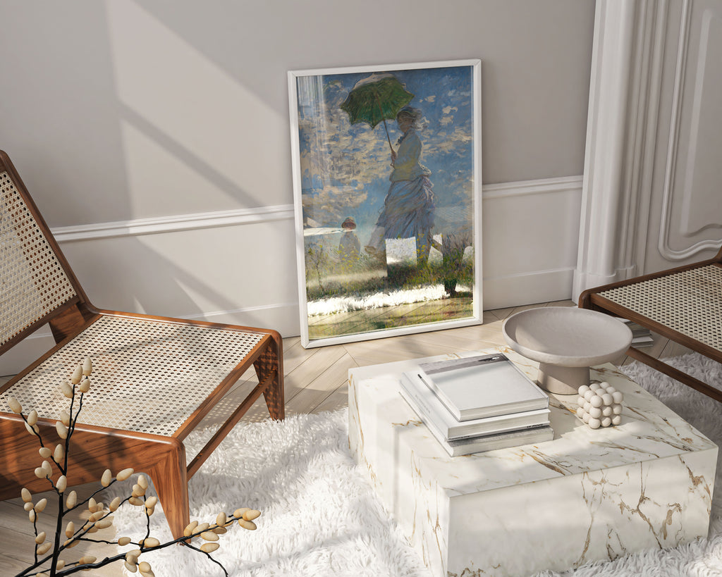 A framed art print of Claude Monet's Impressionist painting, "Woman with a Parasol," leaning against a wall in a modern living room. The wall art is positioned next to two rattan chairs, a marble coffee table, and a fluffy white rug, showcasing how the classic artwork can be integrated into contemporary home decor.