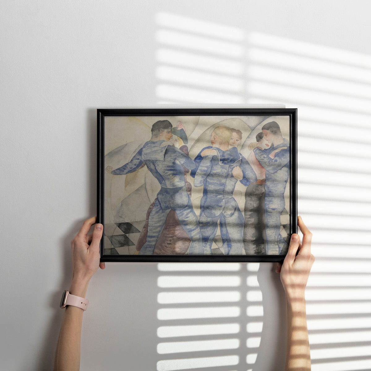 A person holds a black-framed art print against a light gray wall, with soft window-blind shadows cast across the artwork and wall. The print depicts three figures in blue clothing in a muted, painterly style.