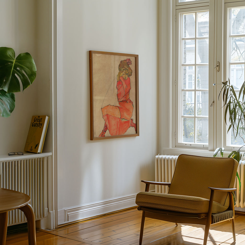 A framed Egon Schiele art print depicting a seated woman in a red dress, hanging on a white wall in a bright room with a wooden chair, window, and plants.