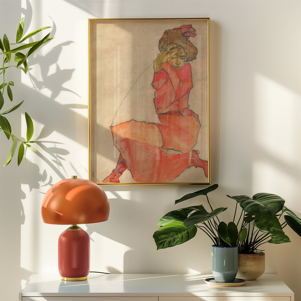An Egon Schiele framed art print featuring a stylised figure in a red dress, hung on a white wall above a console table with a terracotta lamp and potted plants.