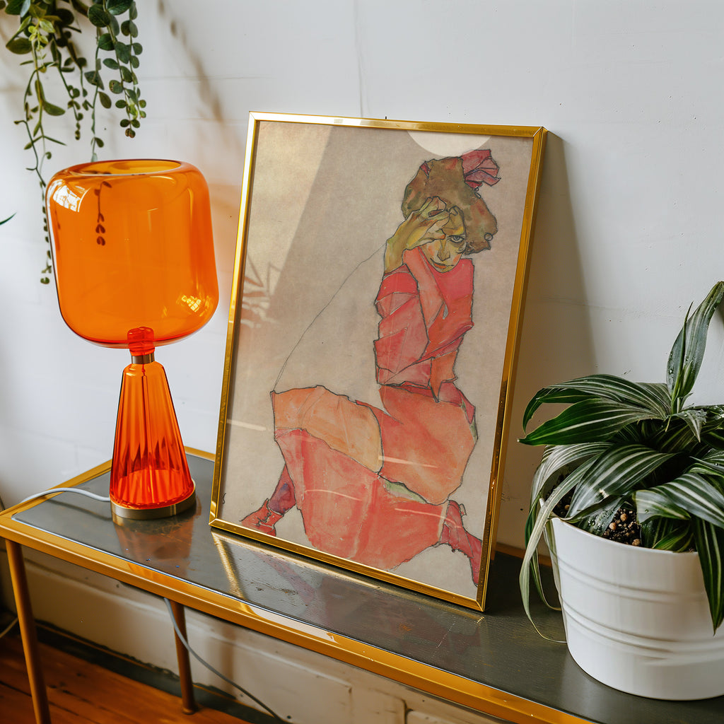 A framed Egon Schiele art print featuring a stylised illustration of a woman in a red dress, displayed on a glass-topped side table next to an amber glass lamp and a potted plant.