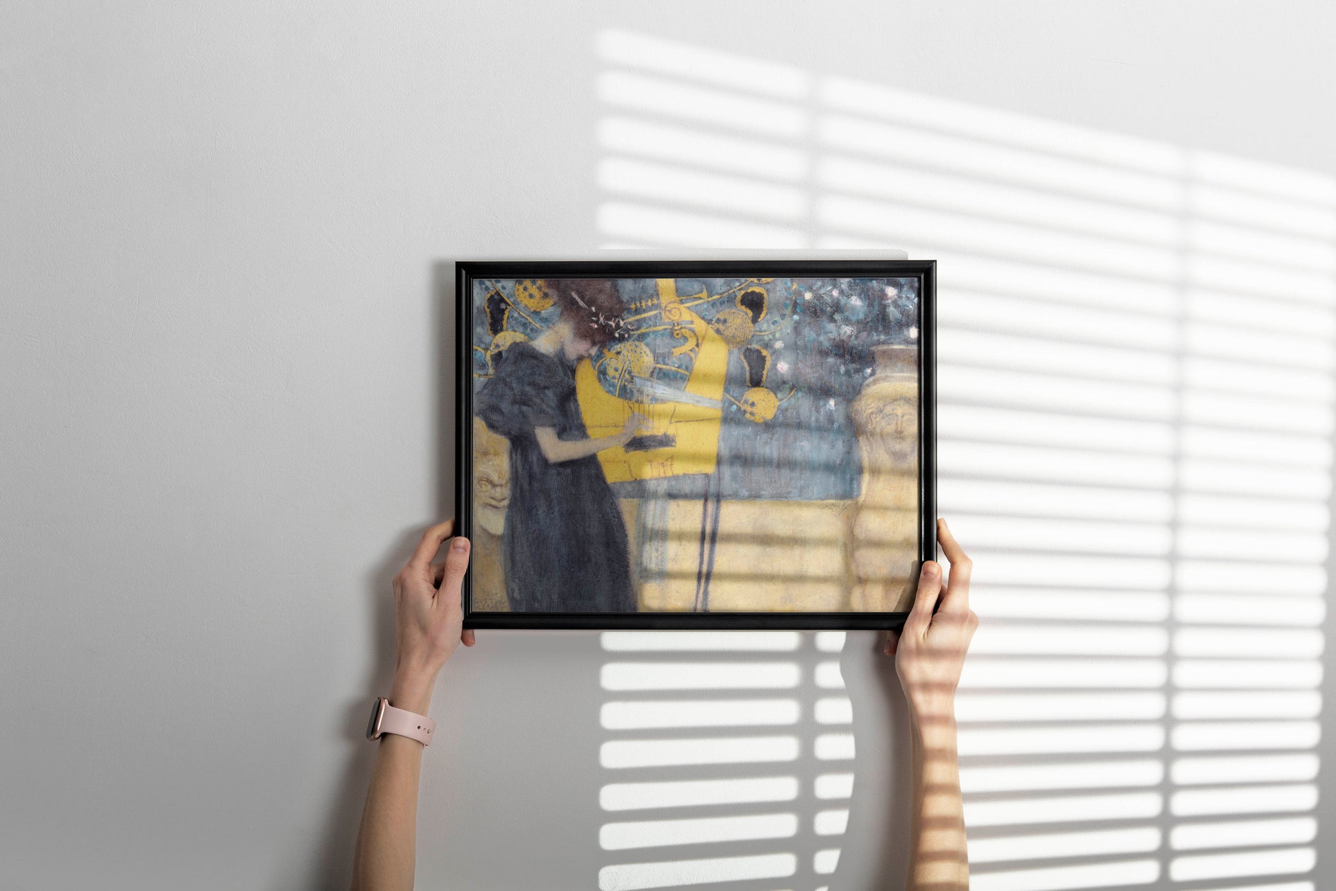 A person's hands holding a black-framed art print of Gustav Klimt's "Music" against a white wall with horizontal blinds casting shadows, showcasing it as stylish home decor.