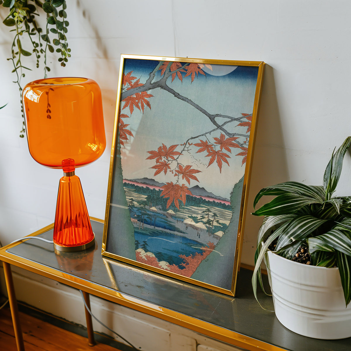A framed art print featuring a Japanese-style landscape with red maple leaves, blue water, and mountains, styled on a gold-framed glass table next to an orange glass lamp and a potted plant.