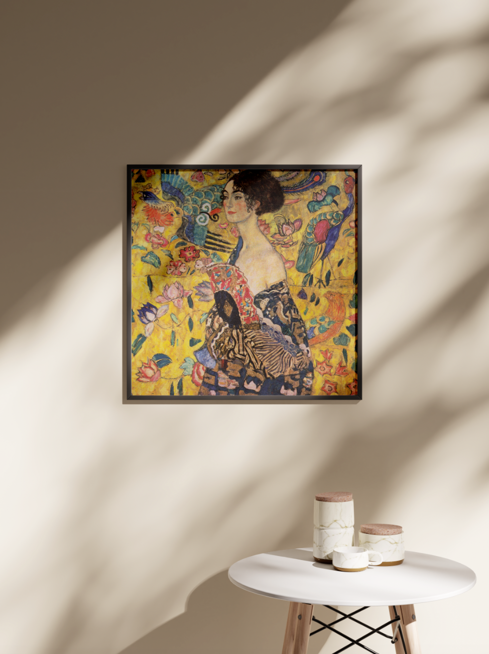 A framed Gustav Klimt "Lady with Fan" giclée art print hanging on a beige wall, with natural light and a shadow of a leaf creating a sophisticated atmosphere above a small white side table.
