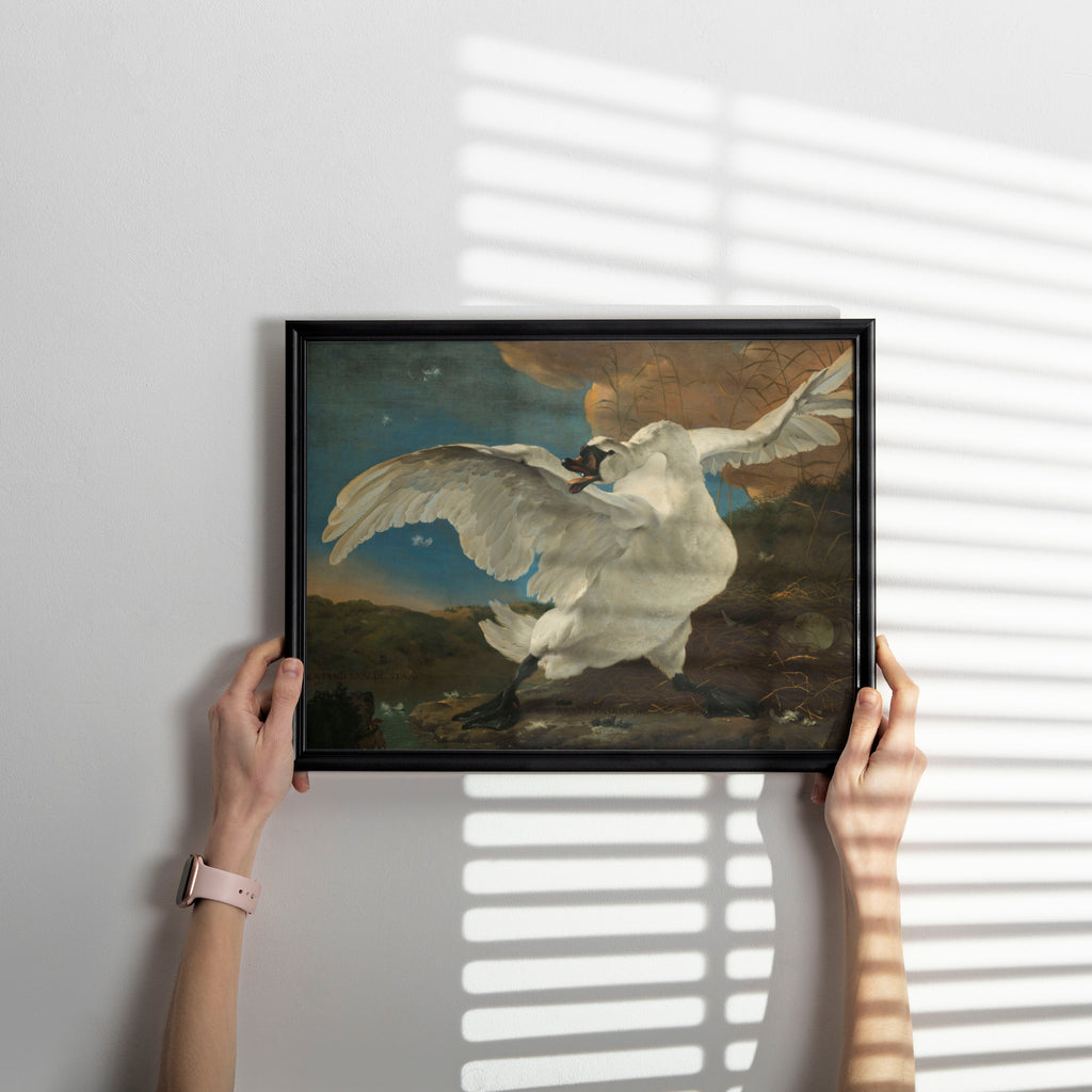 A person holds a black-framed art print depicting a white swan in flight against a dark, moody background, set against a light gray wall with soft window-blind shadows.
