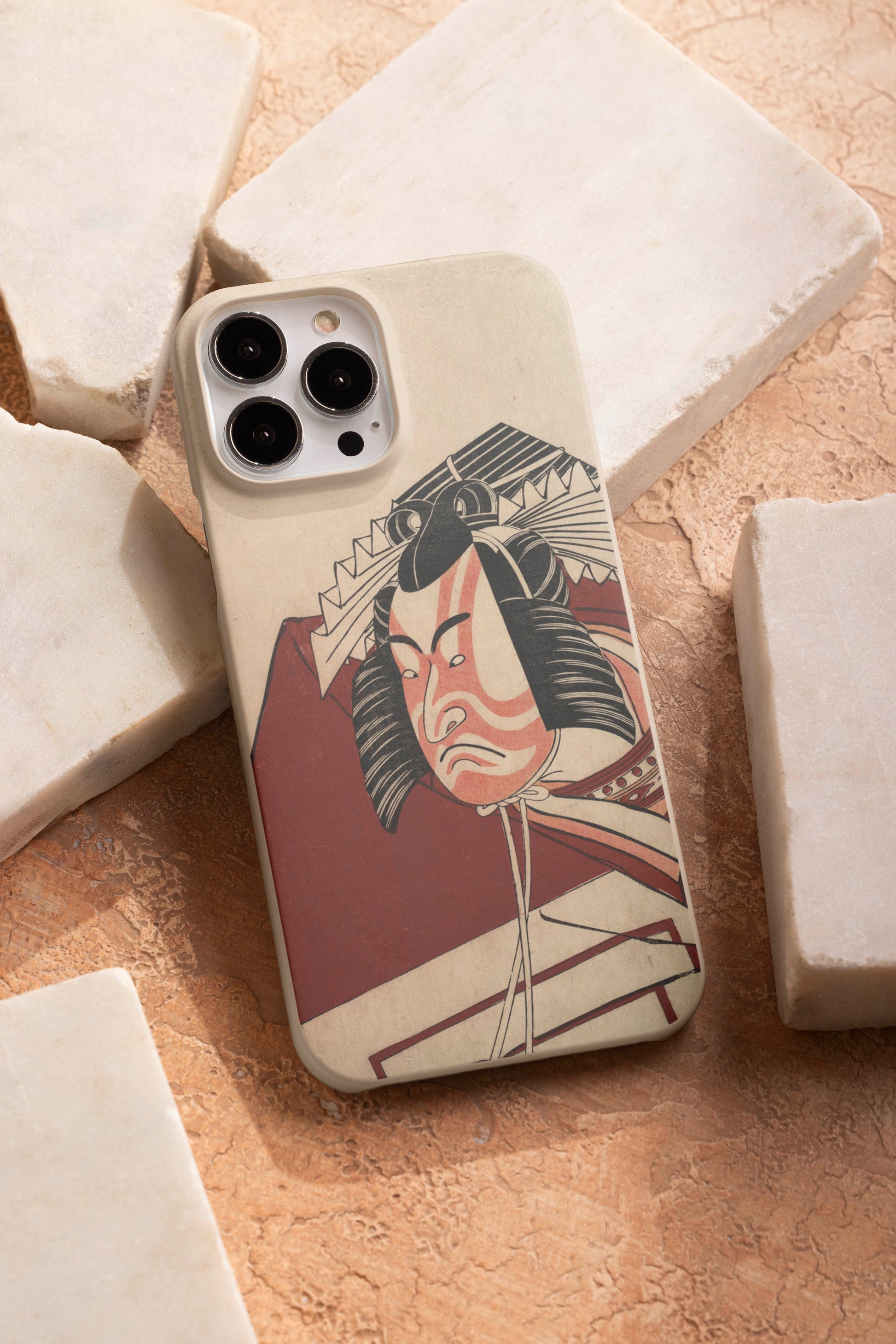 A phone case featuring an ukiyo-e woodblock print of the kabuki actor Ichikawa Monnosuke II, resting on light-coloured stone slabs. The phone case has a textured, off-white background and the print shows the actor with dramatic red and white makeup and an elaborate wig.