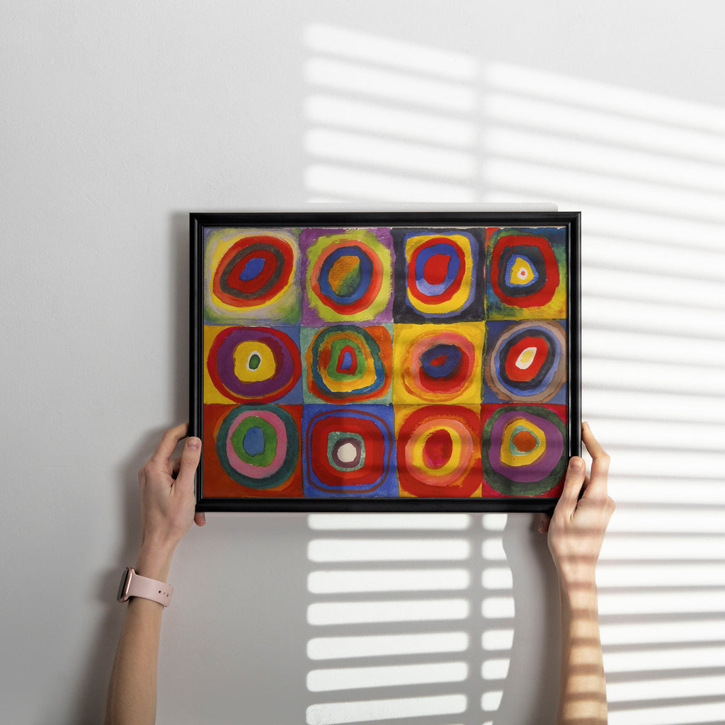 A person holds a black-framed abstract art print featuring a 3x3 grid of colourful concentric circle and square patterns in red, yellow, blue, green, and purple, set against a light gray wall with soft window-blind shadows.