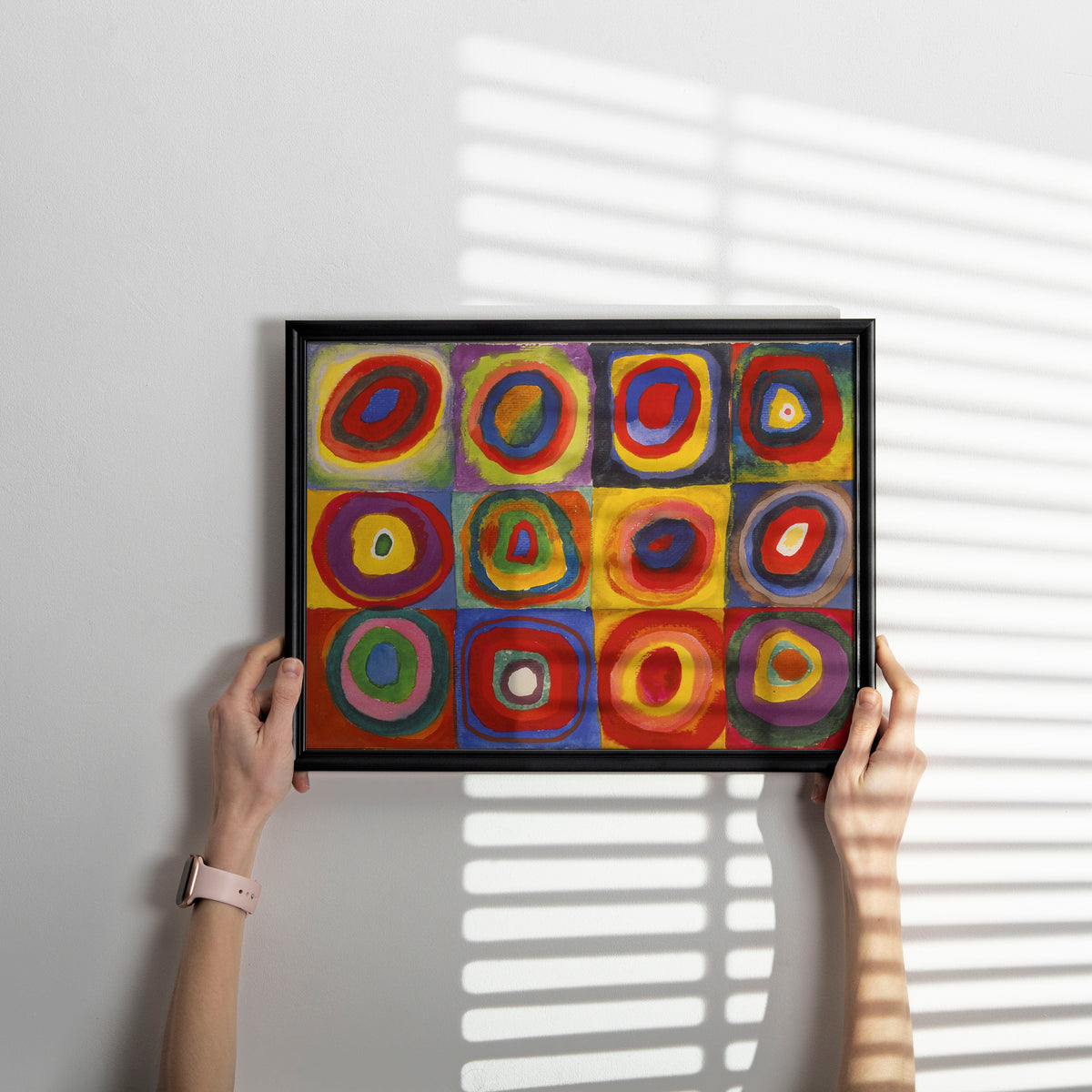 A person holds a black-framed abstract art print featuring a 3x3 grid of colourful concentric circle and square patterns in red, yellow, blue, green, and purple, set against a light gray wall with soft window-blind shadows.