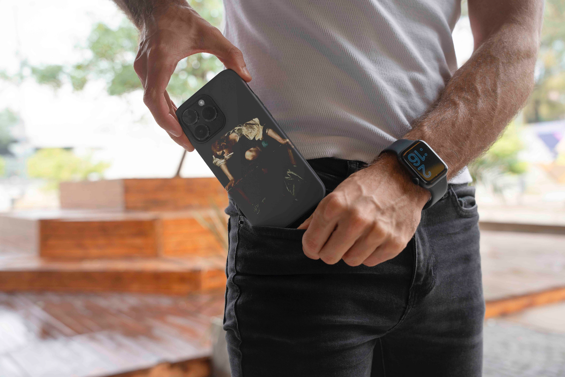 A man in black jeans and a white t-shirt putting a black phone case, featuring Caravaggio's Narcissus painting, into his front pocket. He is wearing a black smartwatch.