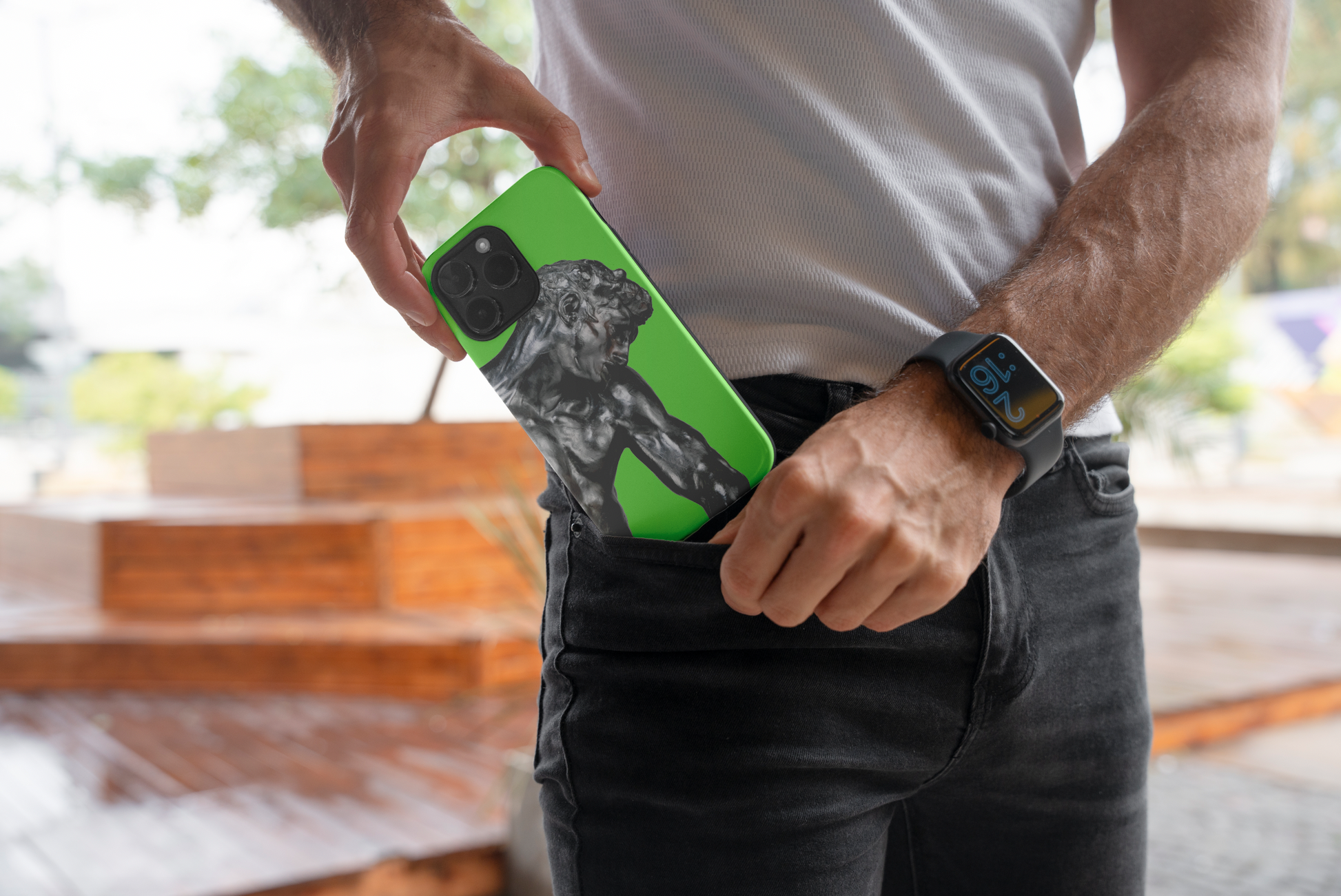 A man putting a green phone case featuring Auguste Rodin's "Adam" sculpture into the back pocket of his dark jeans. He is wearing a white t-shirt and an Apple Watch. The background is a slightly blurred outdoor setting with a wooden staircase visible.