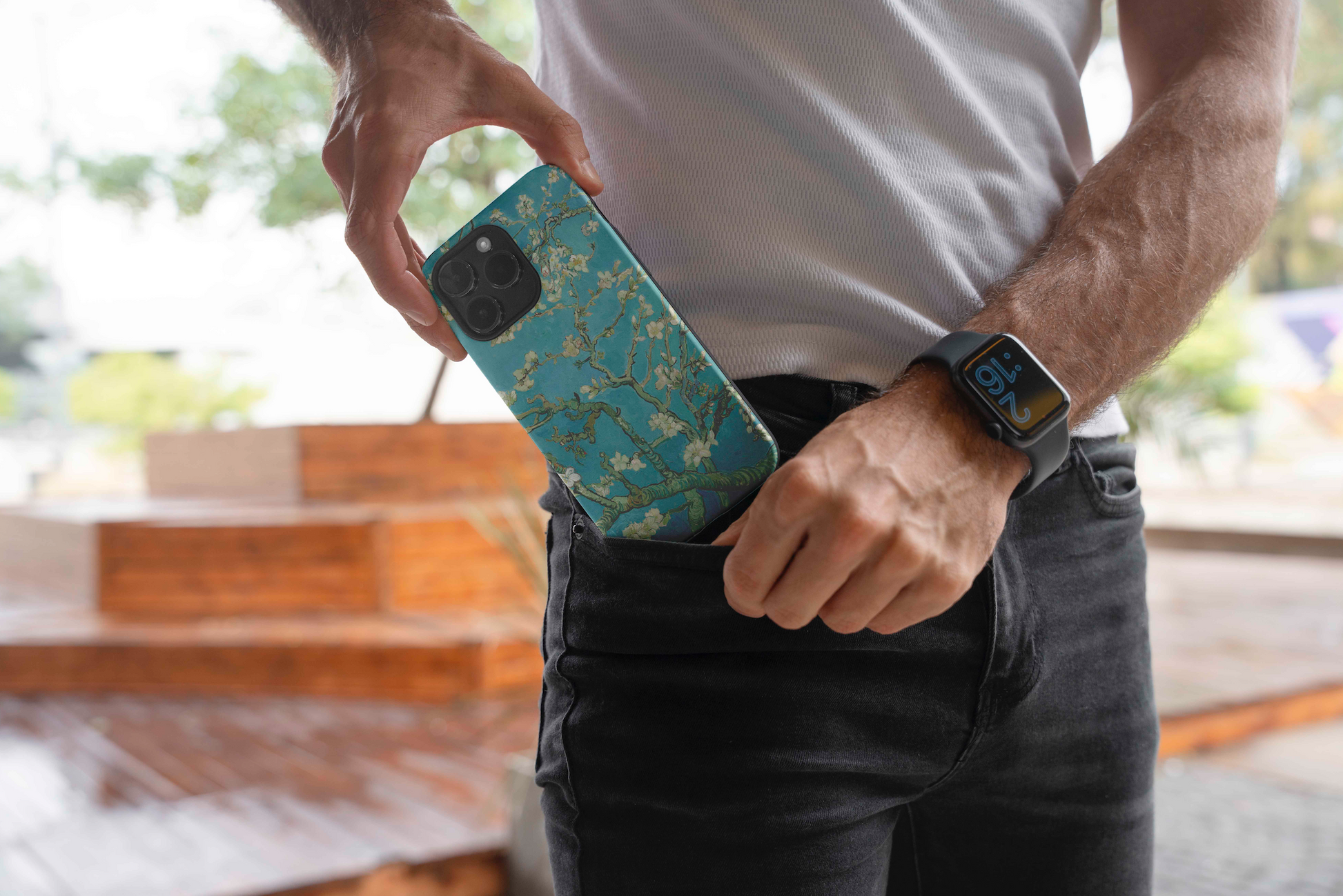 A man in a white t-shirt and dark jeans is seen from the waist down, putting an iPhone with the Van Gogh Almond Blossom case into his front jeans pocket. He is wearing a dark smartwatch on his left wrist.