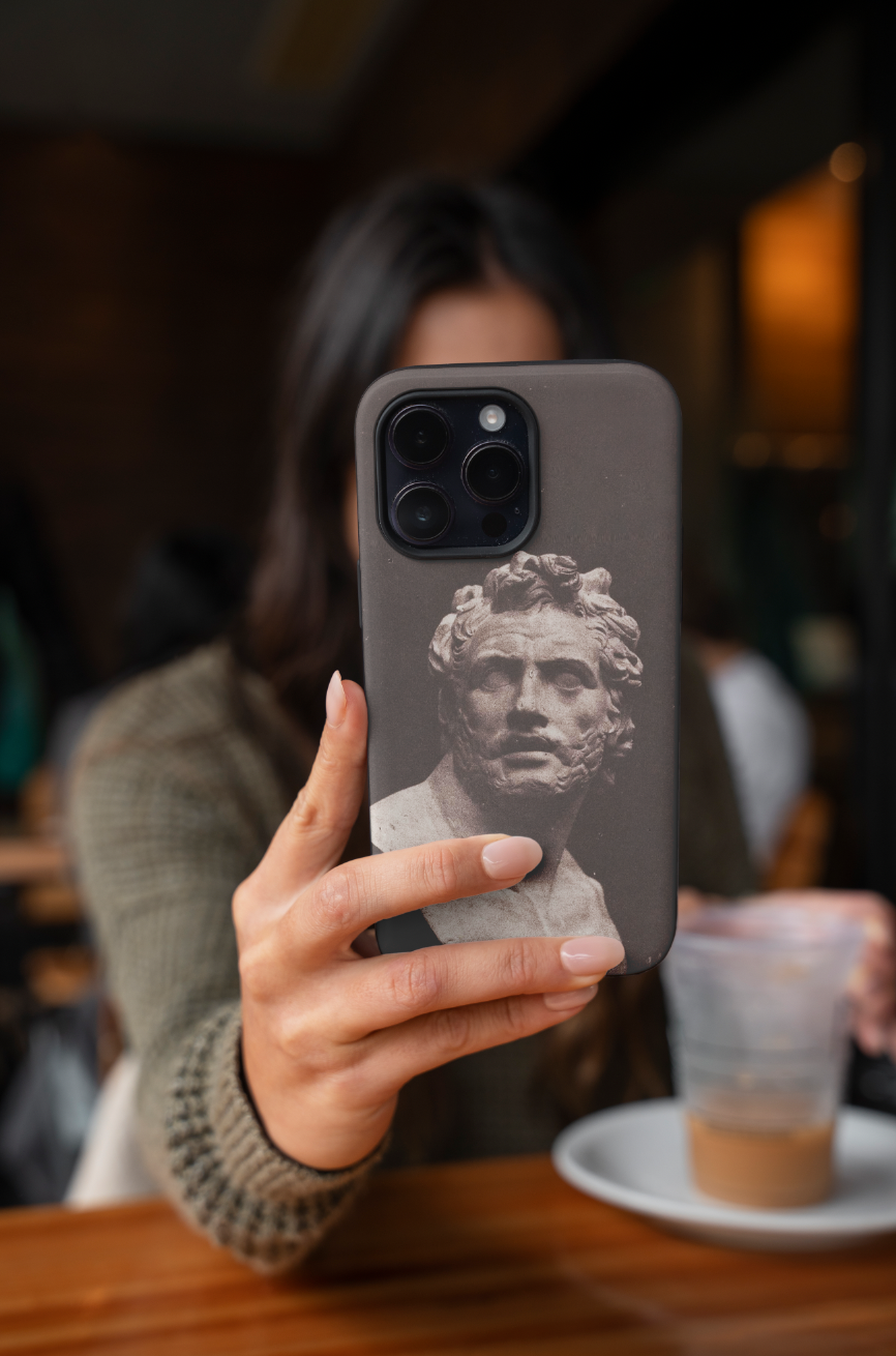A person in a café holding an iPhone with the ART SNOB 'Bust of Patroclus' fine art phone case, showcasing the historical photography print and the elegant design in a lifestyle setting.