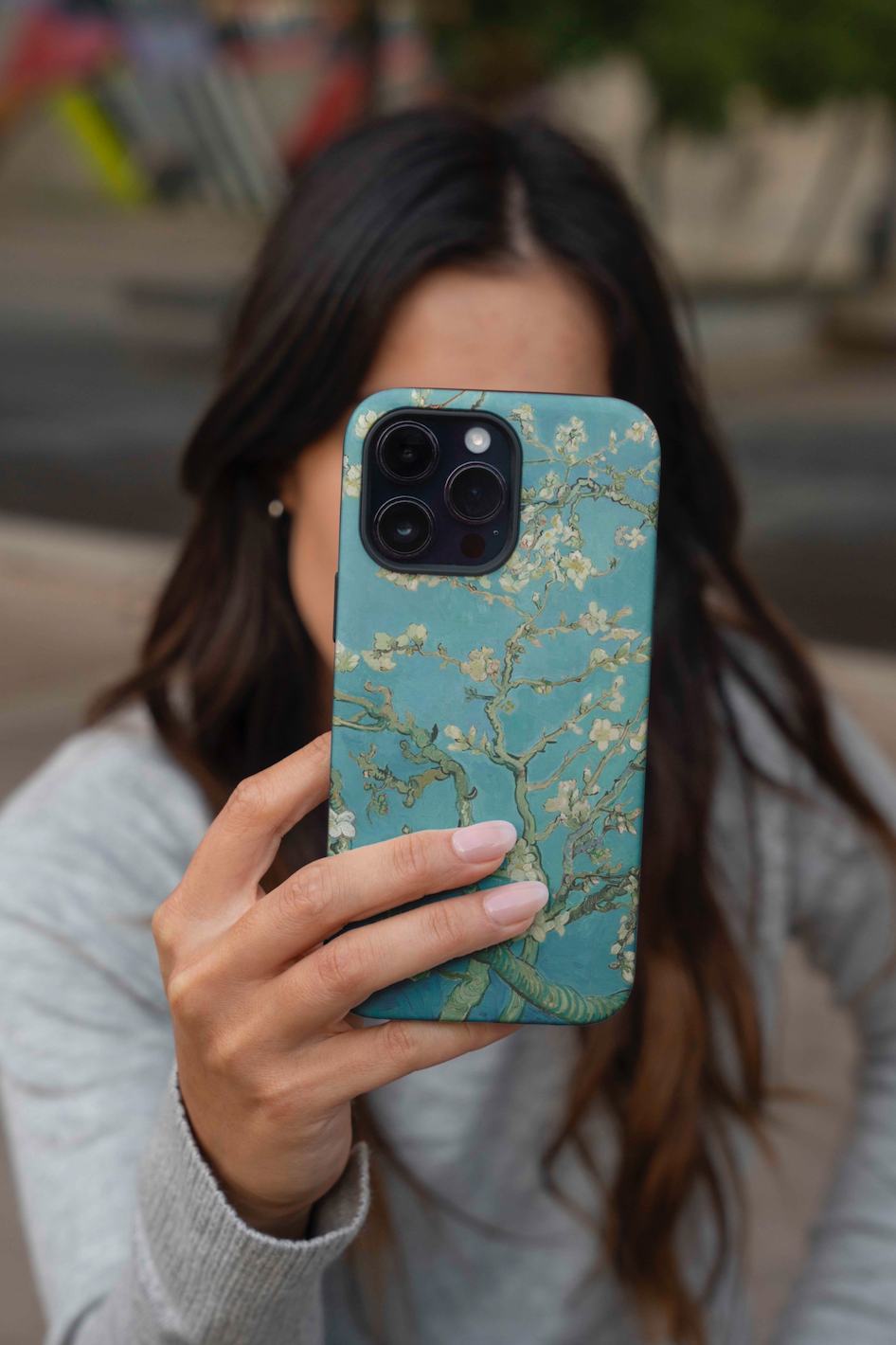 A person with long dark hair holds up an iPhone with the Vincent van Gogh Almond Blossom phone case, obscuring their face. The case features the teal background and white blossoming branches of the famous painting.