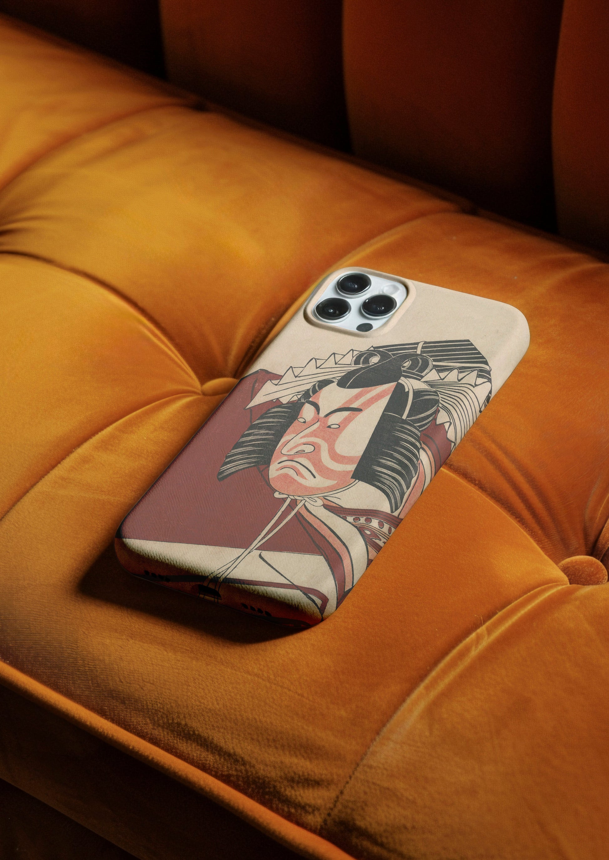 A phone case featuring an ukiyo-e print of a Kabuki actor, resting on the tufted cushions of a dark orange or mustard yellow velvet sofa. The case has a slightly textured, light background and depicts the actor with expressive red and white makeup.