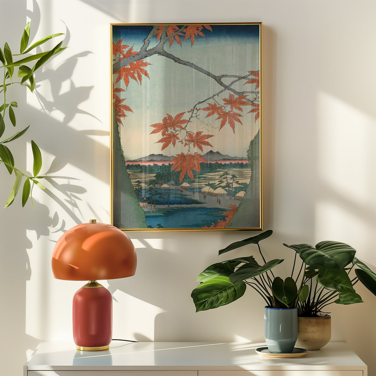 A framed art print featuring a traditional Japanese landscape with red autumn leaves hanging in the foreground, displayed on a white console table in a styled room with a terracotta lamp and potted plants.