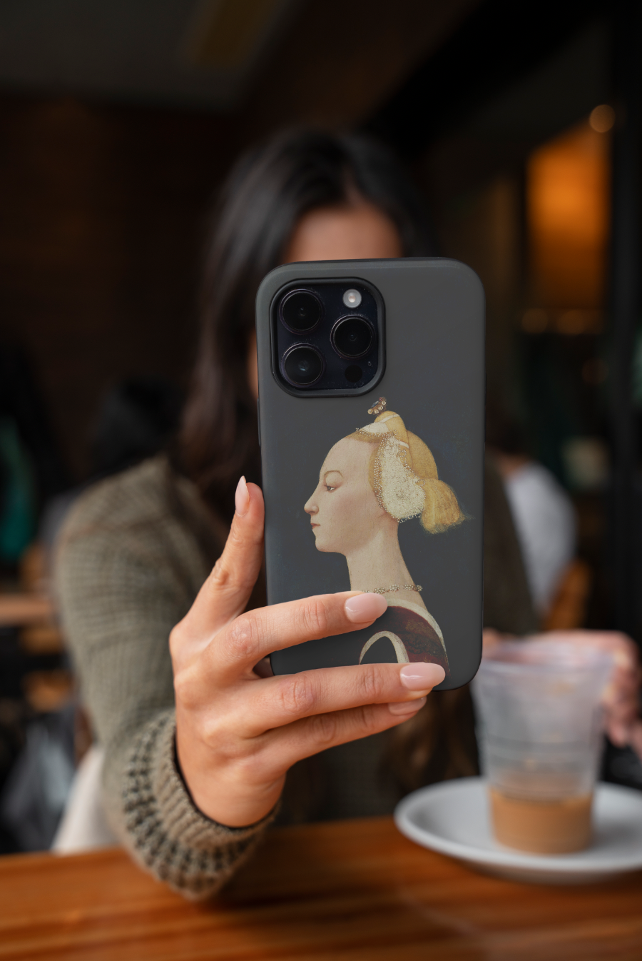 A person holds up a phone with the ART SNOB "Master of the Castello Nativity Portrait of a Woman" phone case in a cafe setting. The focus is on the phone case and the classic art print, with the person and background blurred to highlight the product in a lifestyle context.