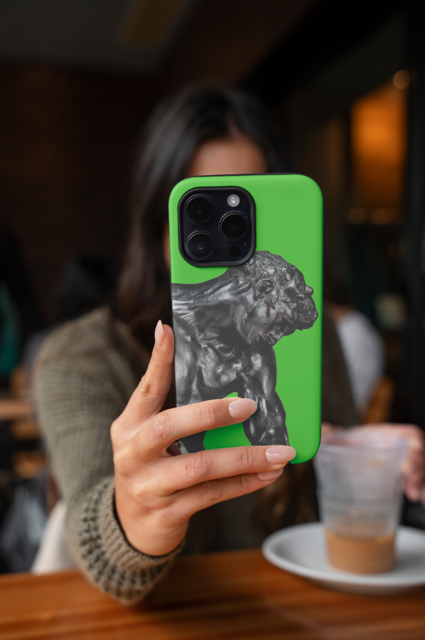 A woman holding up a green phone with a phone case featuring Auguste Rodin's bronze sculpture, "Adam." The woman is seated at a wooden table in what appears to be a cafe, with a coffee cup in a clear plastic cup visible in the foreground. The focus is on the phone case and the woman's hand, while the background is softly blurred.